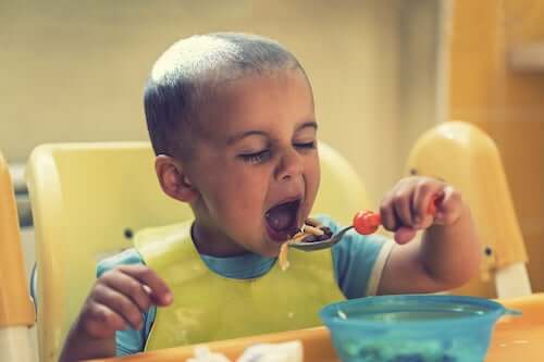 Bambino di due anni seduto sul seggiolone che mangia da solo utilizzando il cucchiaio.